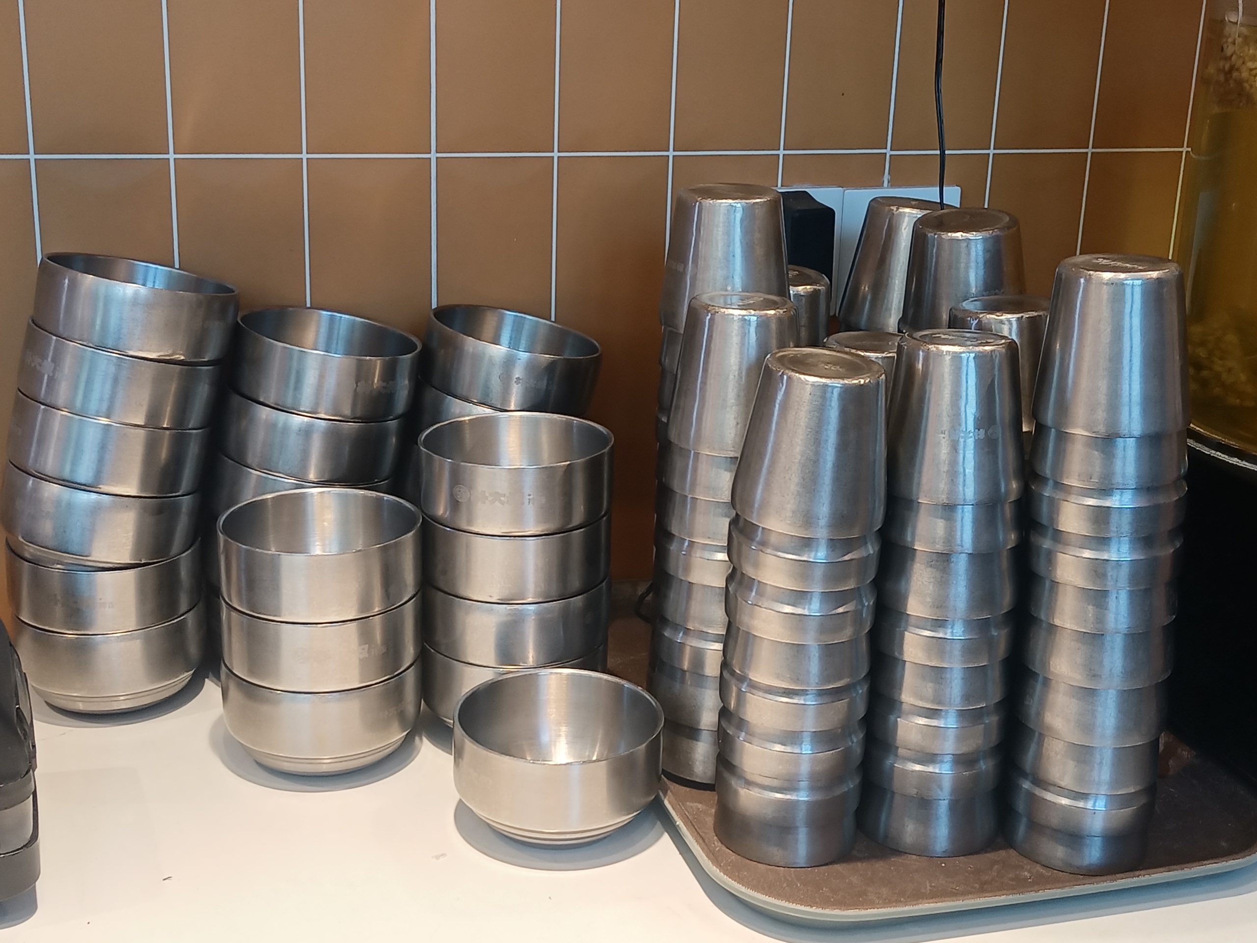 Stacks of stainless steel bowls and cups on a tray against a tiled kitchen wall