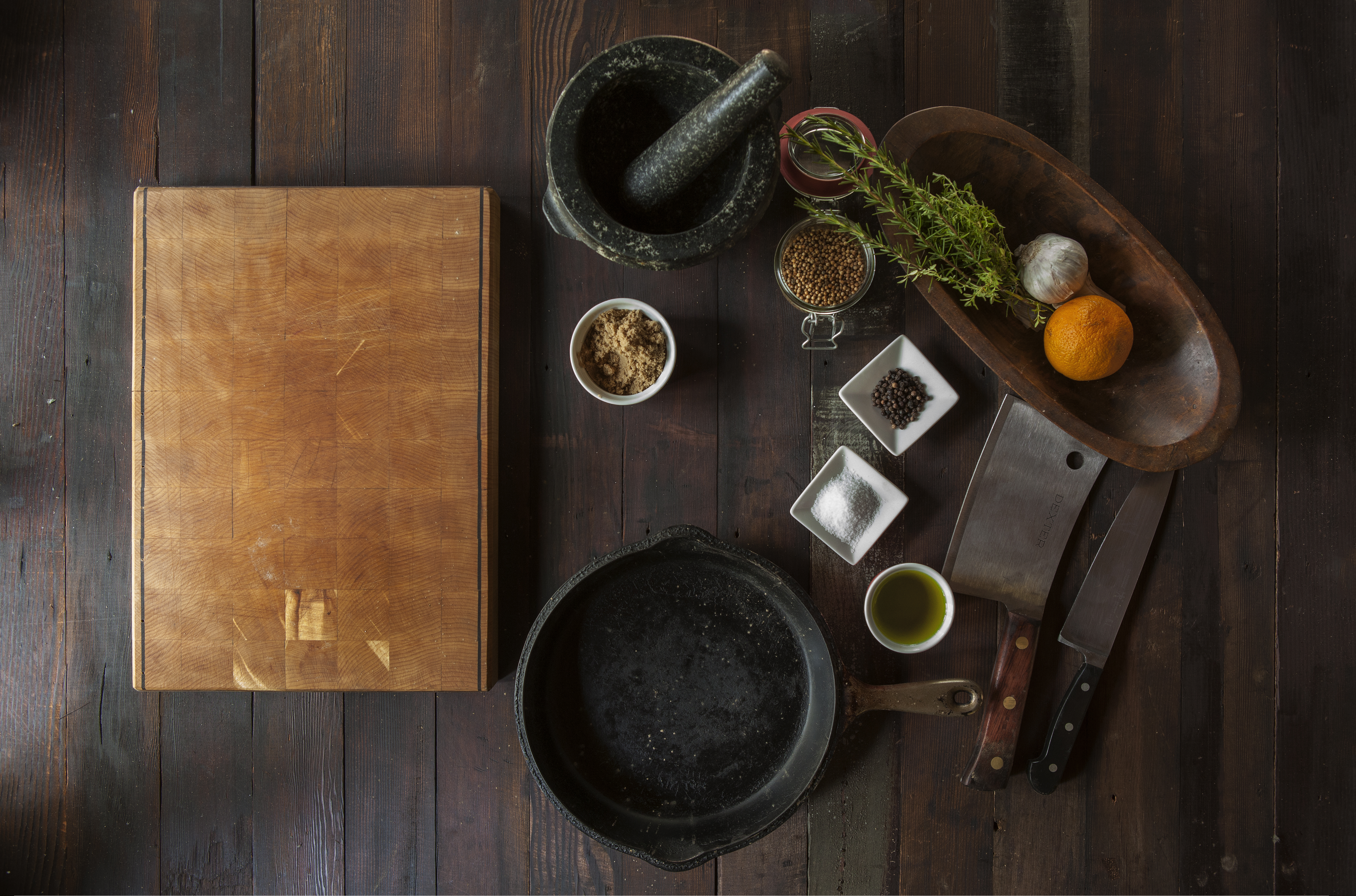Wooden cutting board, cleaver, cast iron pan and small bowls of herbs and spices on a dark table