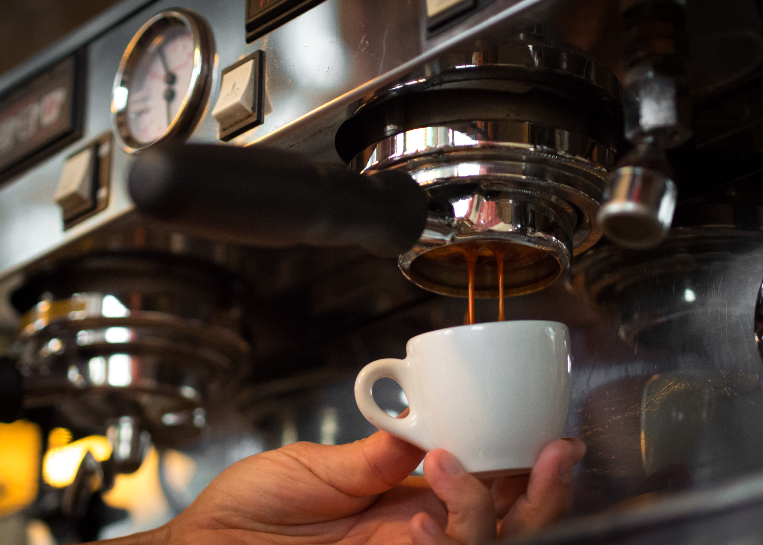 Espresso pouring from a commercial machine into a small white cup held by hand