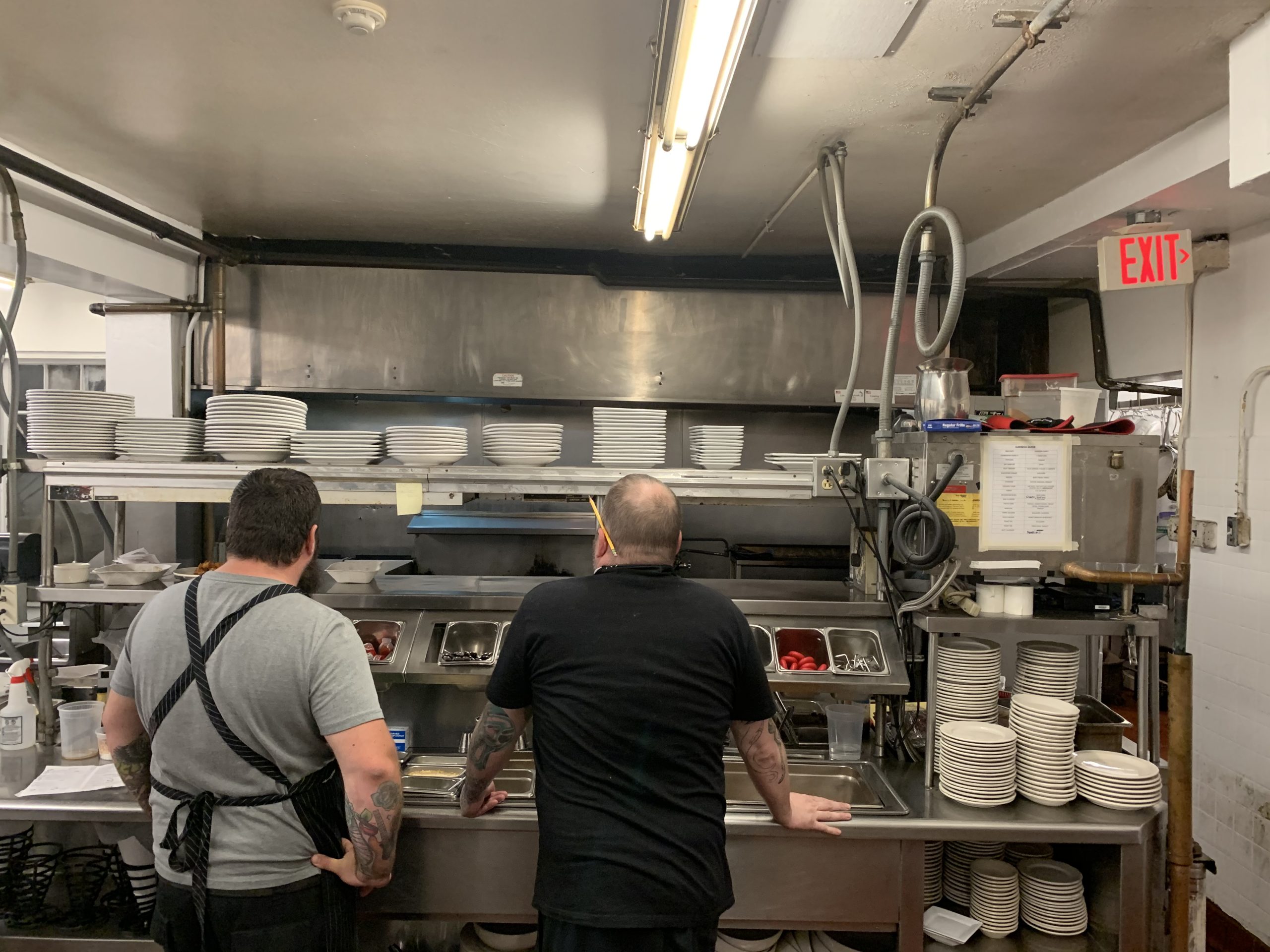 Two cooks seen from behind at a restaurant kitchen expo station with stacked plates