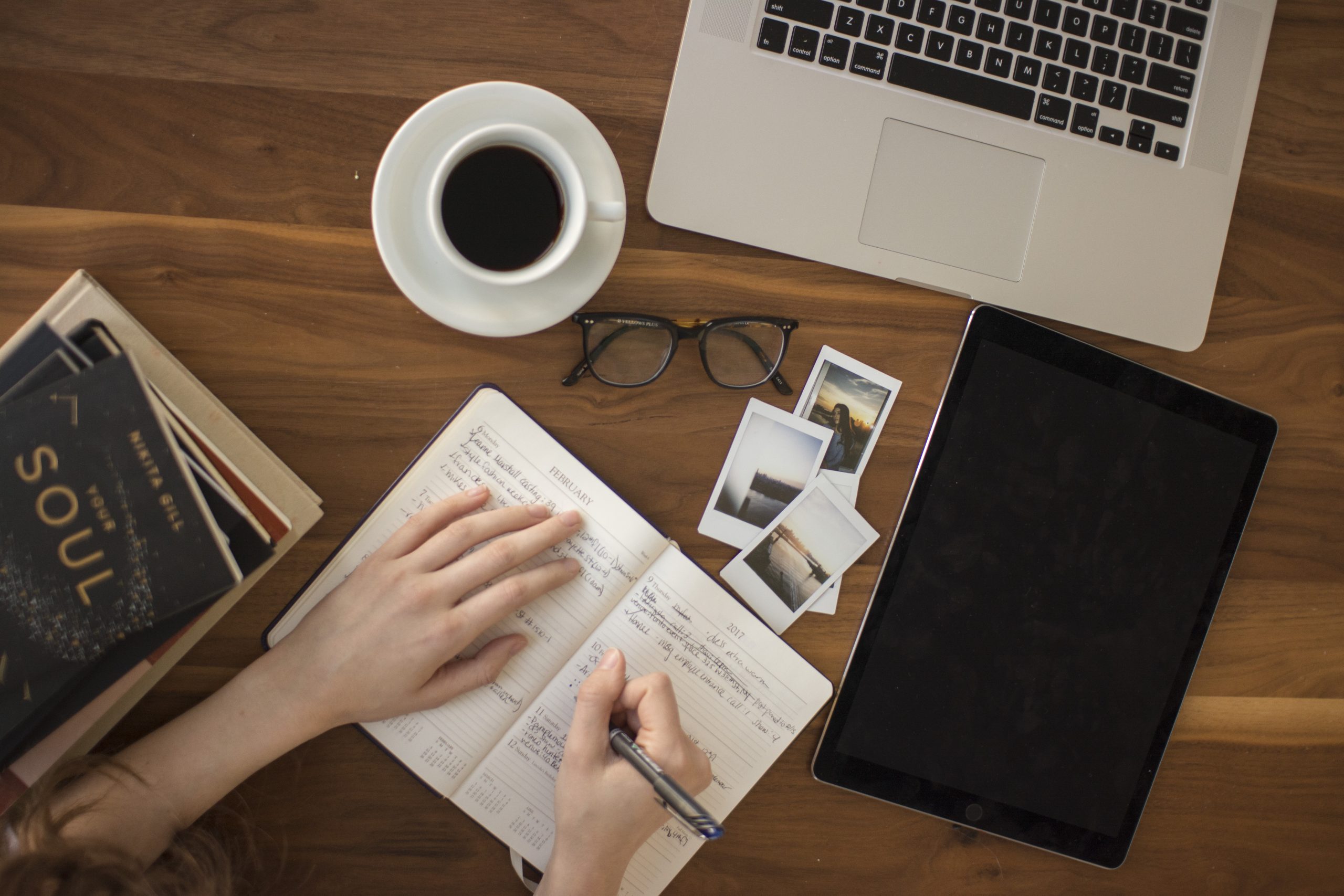 Hand writing in a planner beside a cup of black coffee, glasses and a tablet on a wooden table