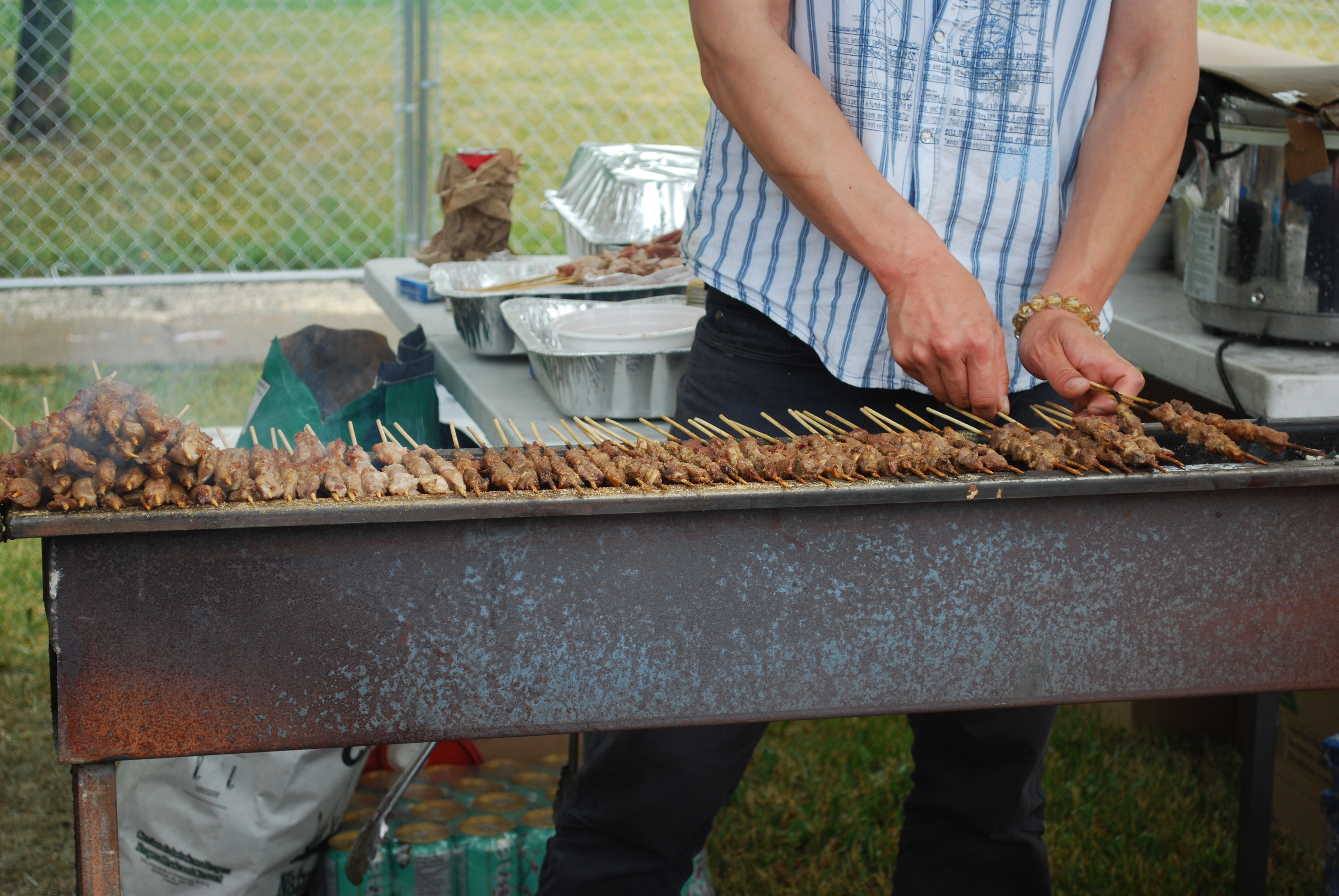 Vendor turning rows of meat skewers on a long outdoor grill at an event