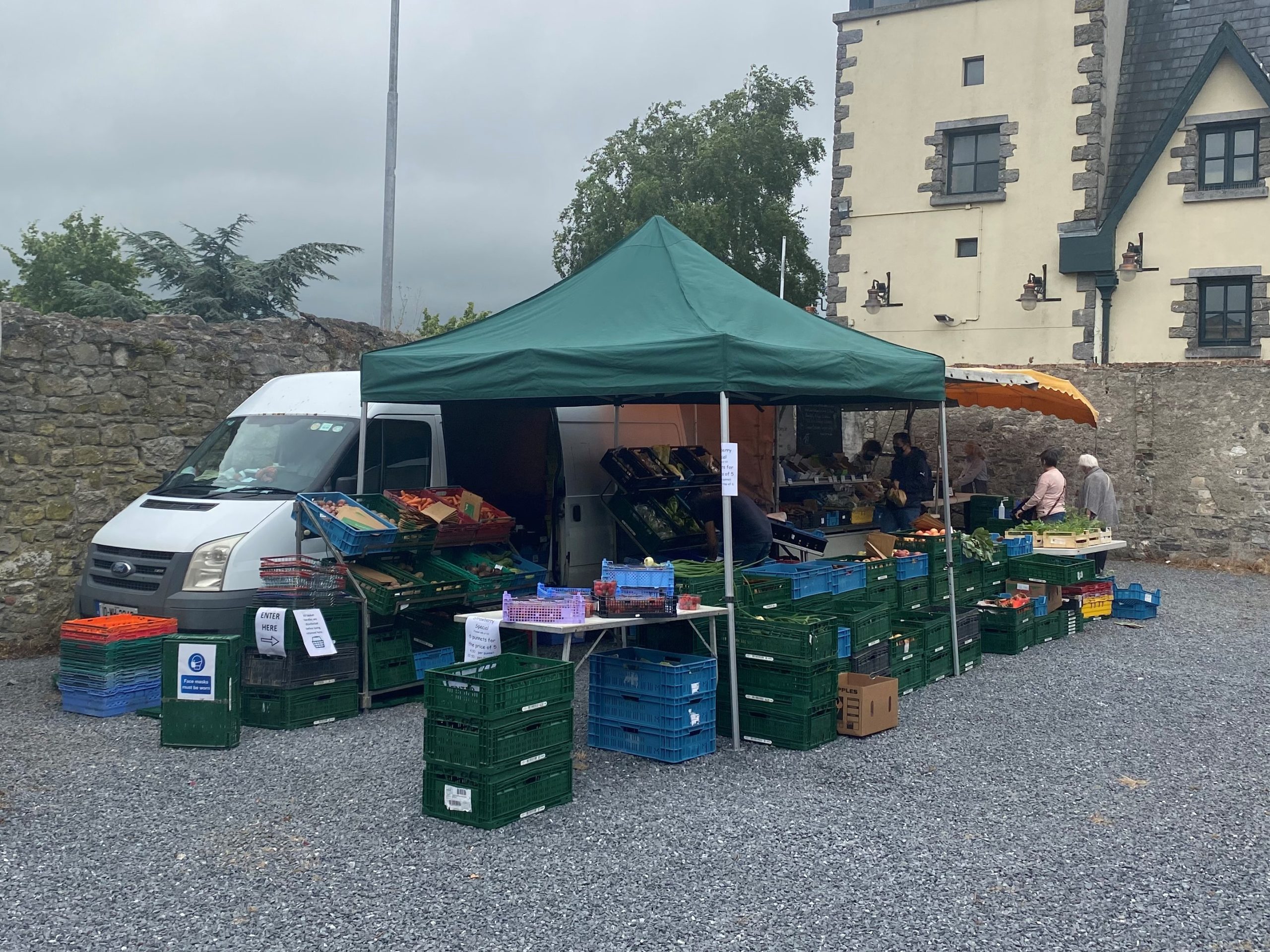Green gazebo market stall with crates of vegetables on a gravel square beside a white van