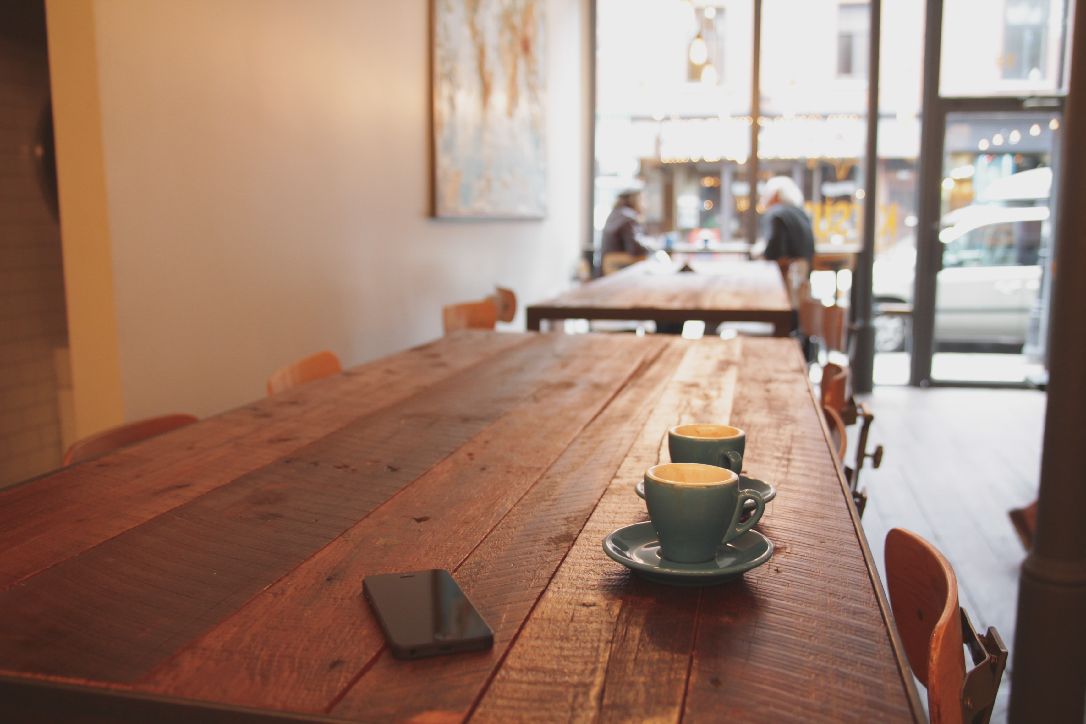 Two espresso cups on a long wooden communal table in a quiet cafe with a large front window