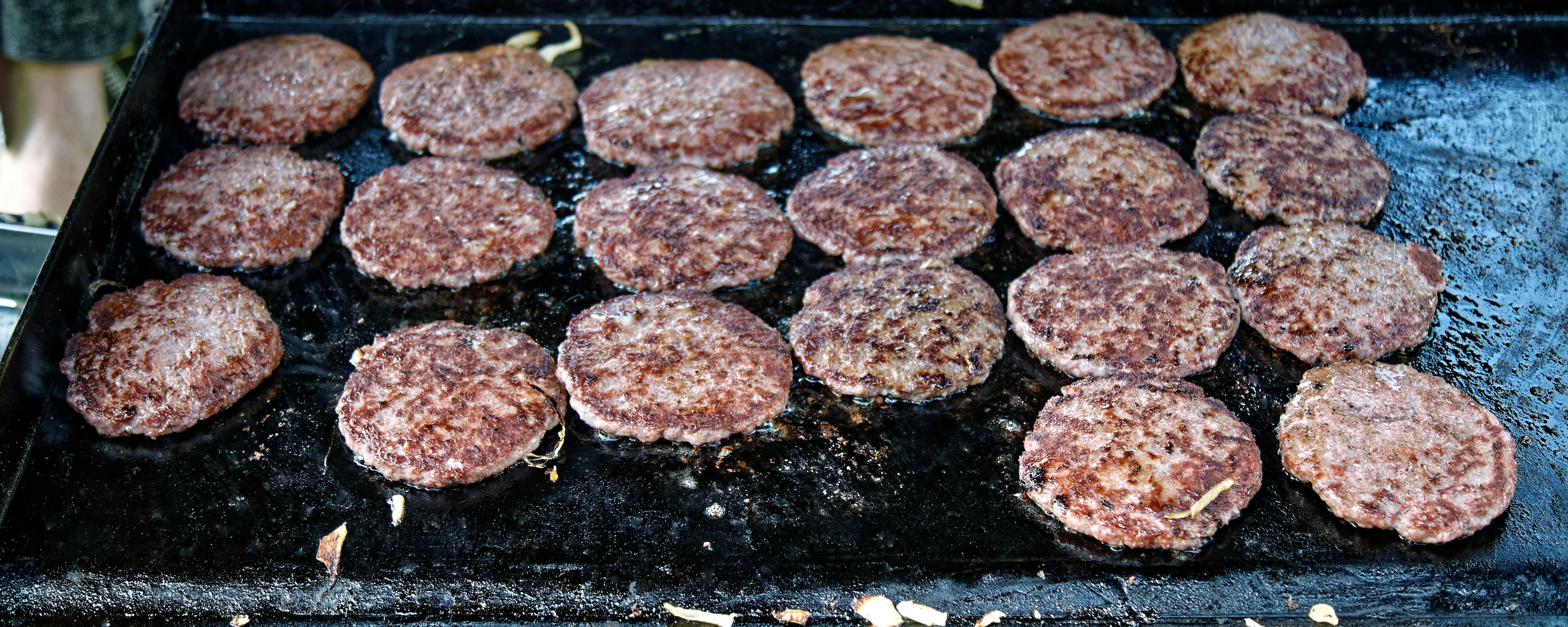 Rows of hamburger patties cooking on a large flat-top griddle at an outdoor event