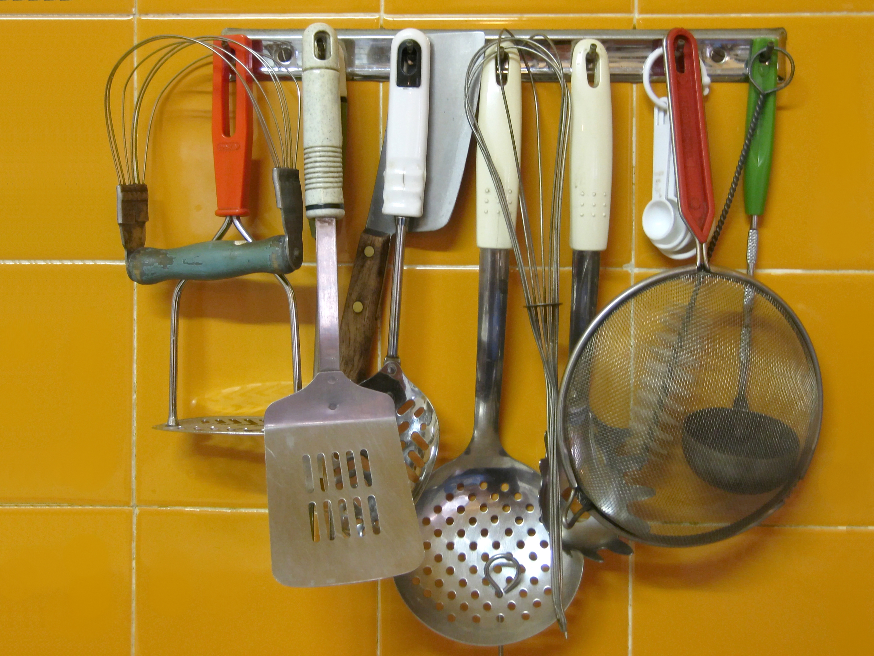 Whisks, a spatula, a slotted spoon and a sieve hanging from a rail on yellow kitchen tiles