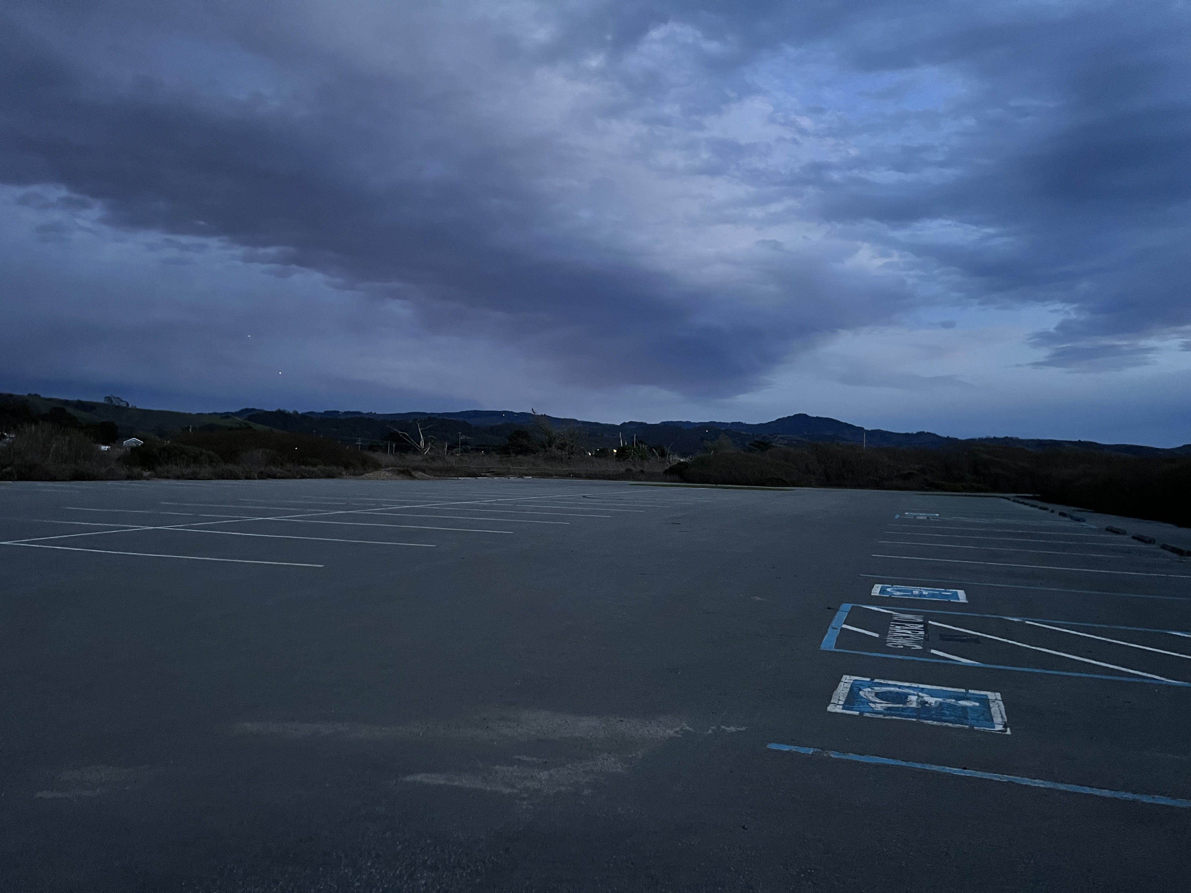 A wide, empty paved parking lot with painted bays under a cloudy evening sky