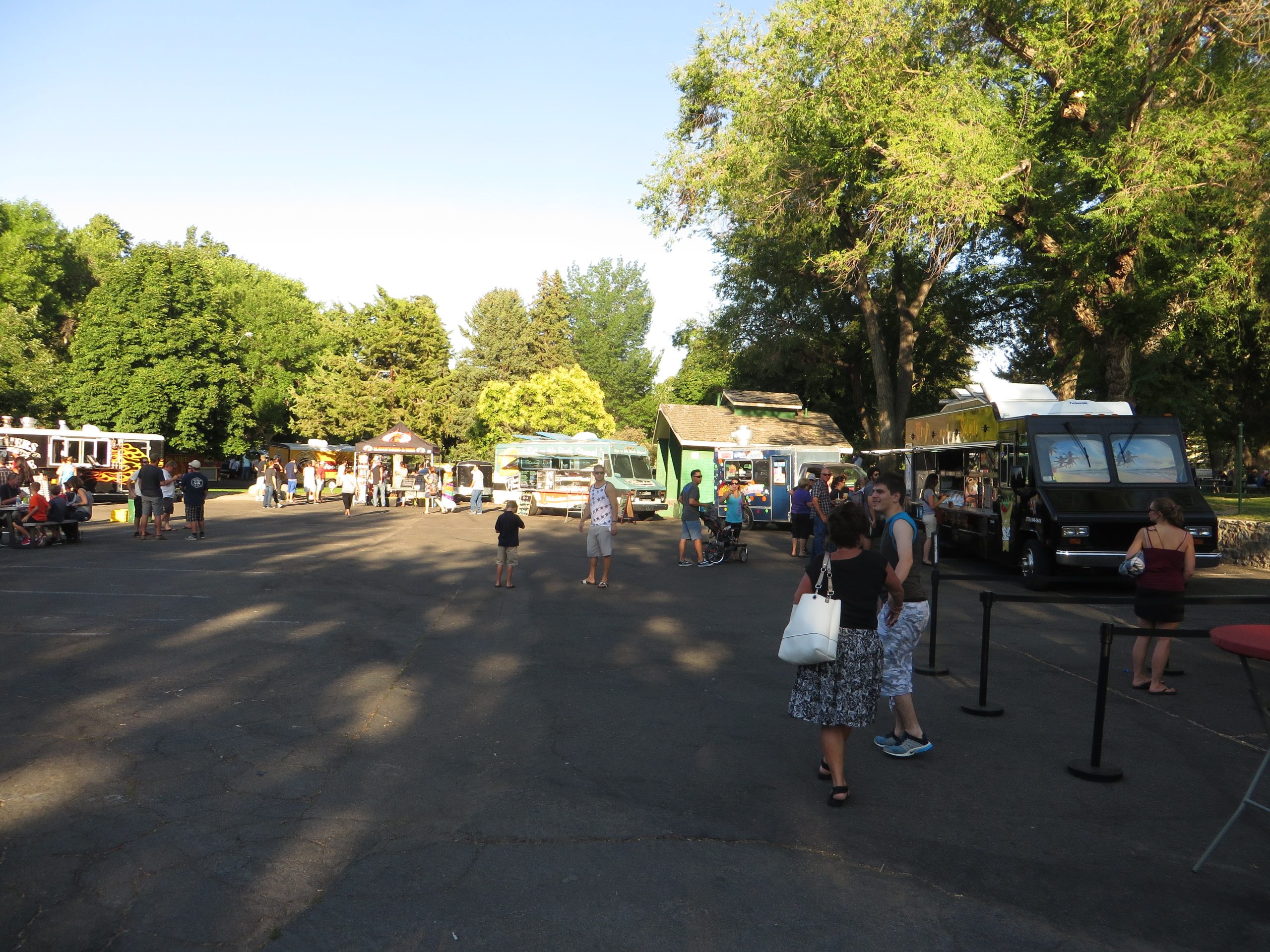 Several food trucks parked around a paved lot in a tree-lined park with people walking between them