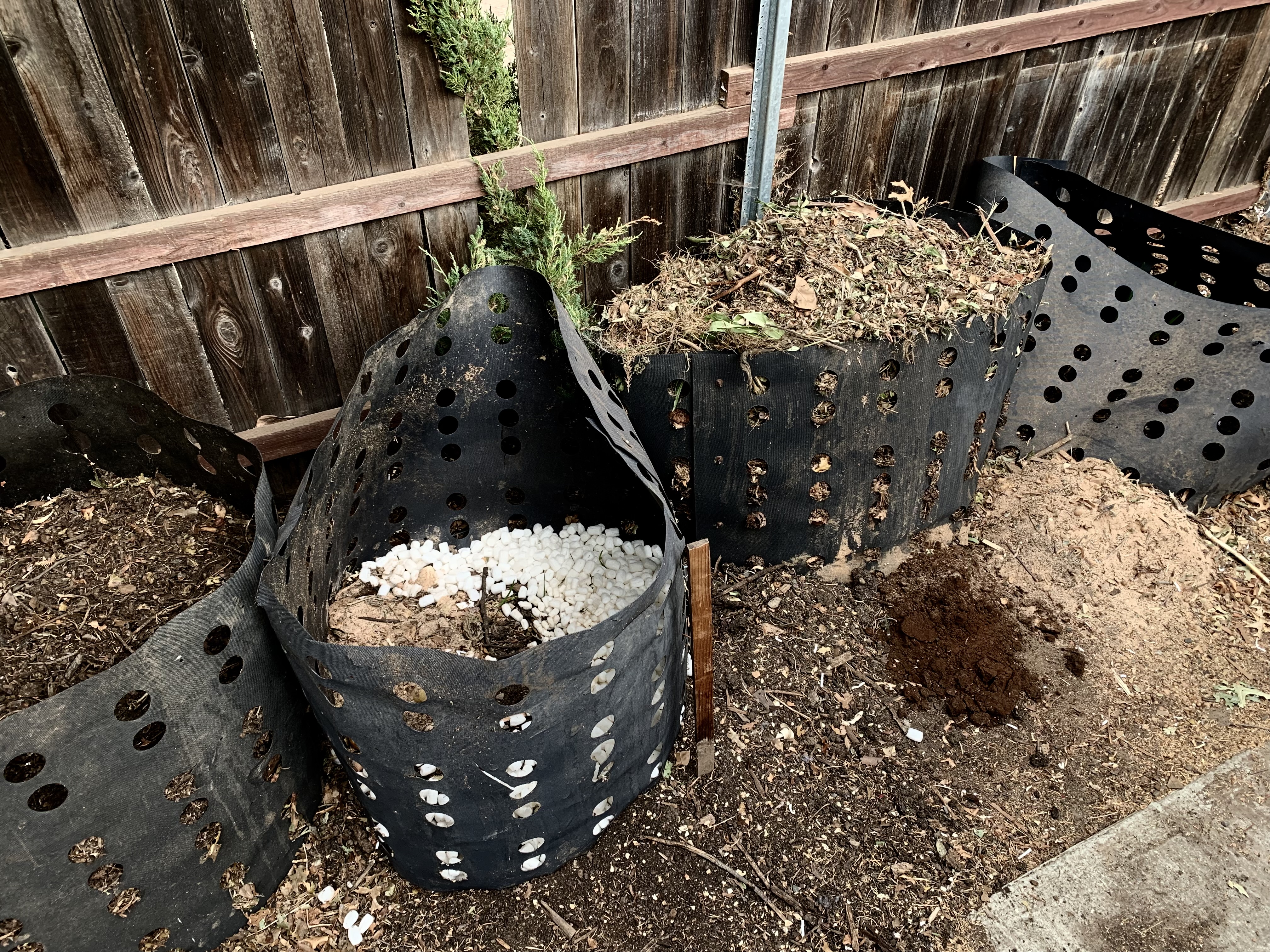 Four black perforated compost bins against a wooden fence, filled with garden and kitchen material