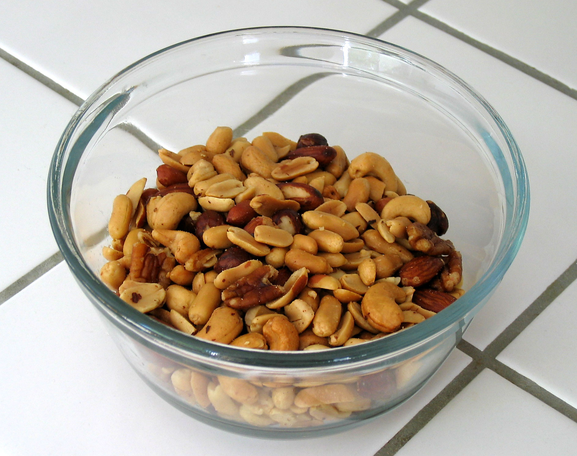 A glass bowl of mixed peanuts, cashews, almonds and pecans on a white tiled counter