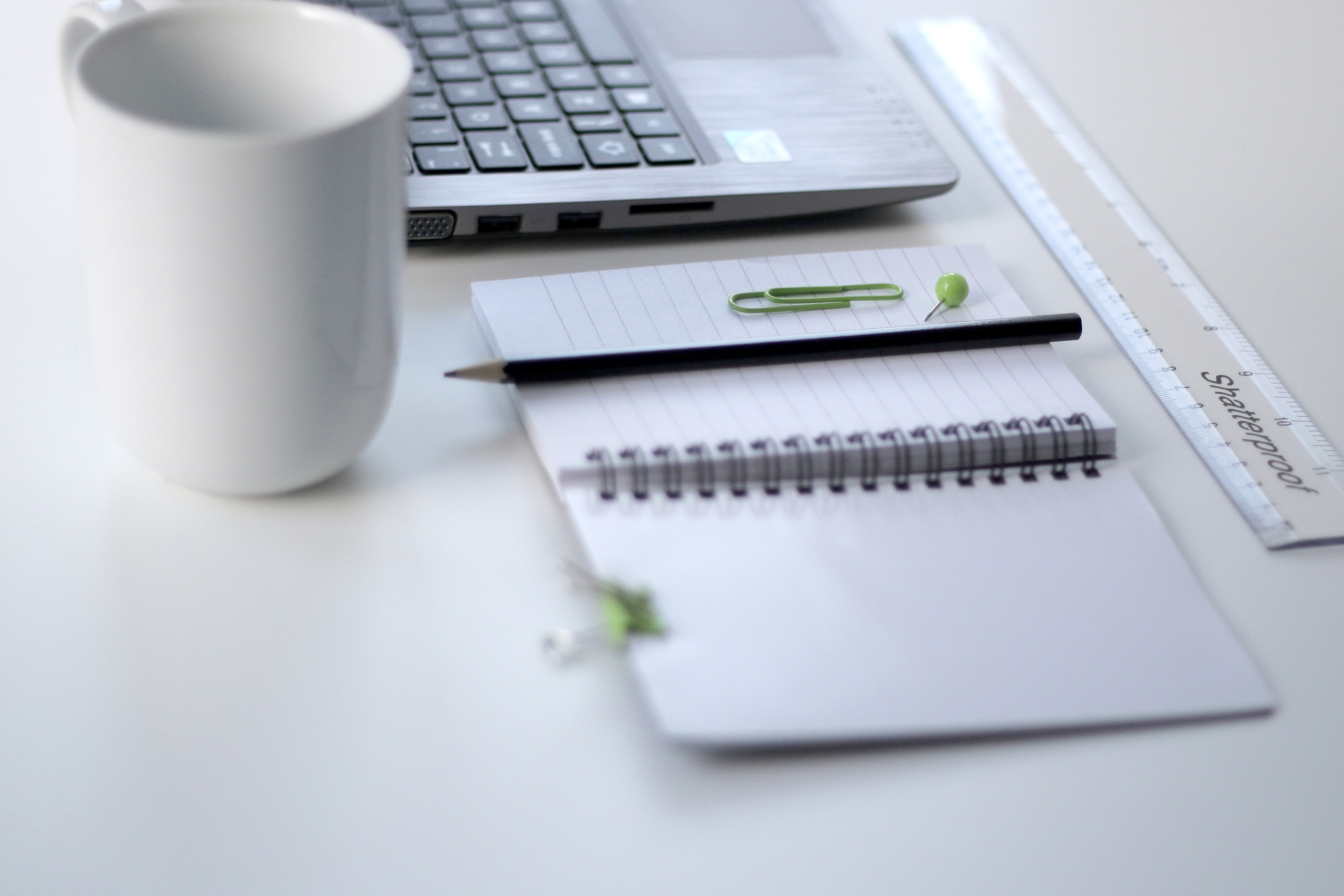 A notebook, pencil, ruler, white mug and laptop on a clean white desk