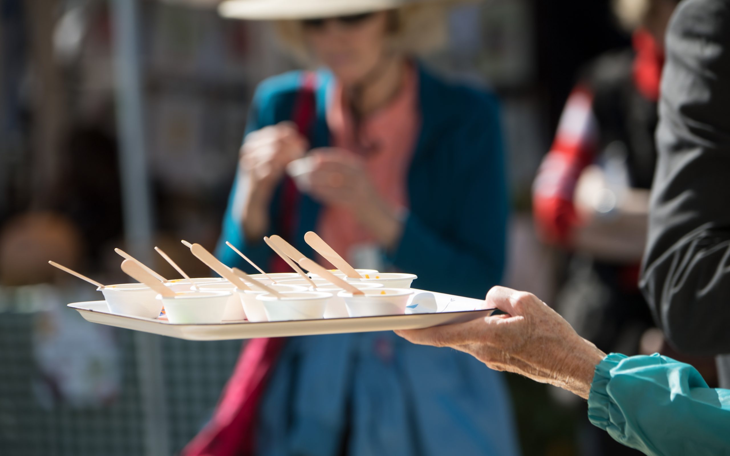 A hand holding a tray of small sample cups with wooden spoons at an outdoor food market