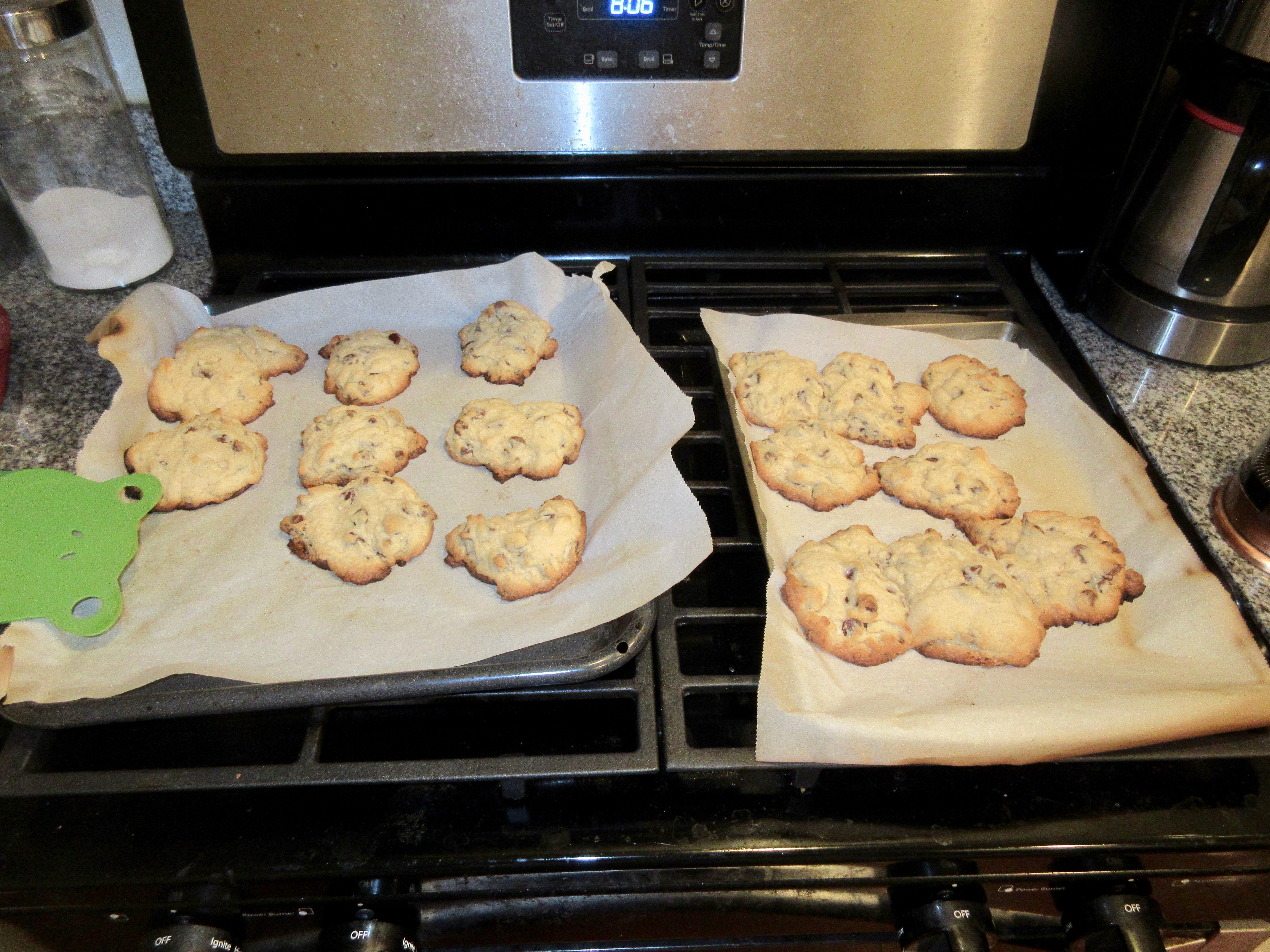 Two trays of freshly baked chocolate chip cookies on parchment resting on a home stovetop