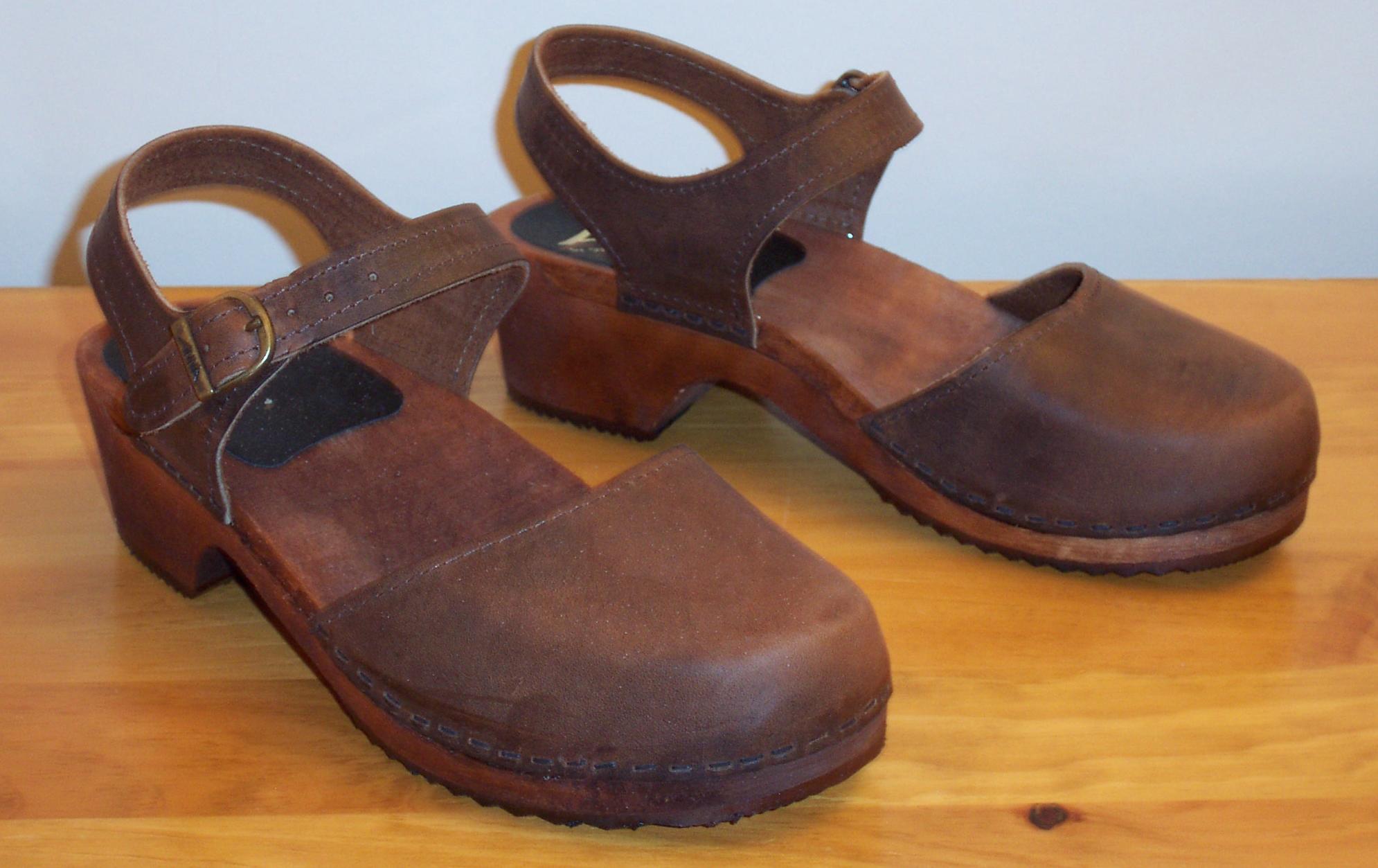 A pair of brown leather clogs with wooden soles and ankle straps on a wooden surface
