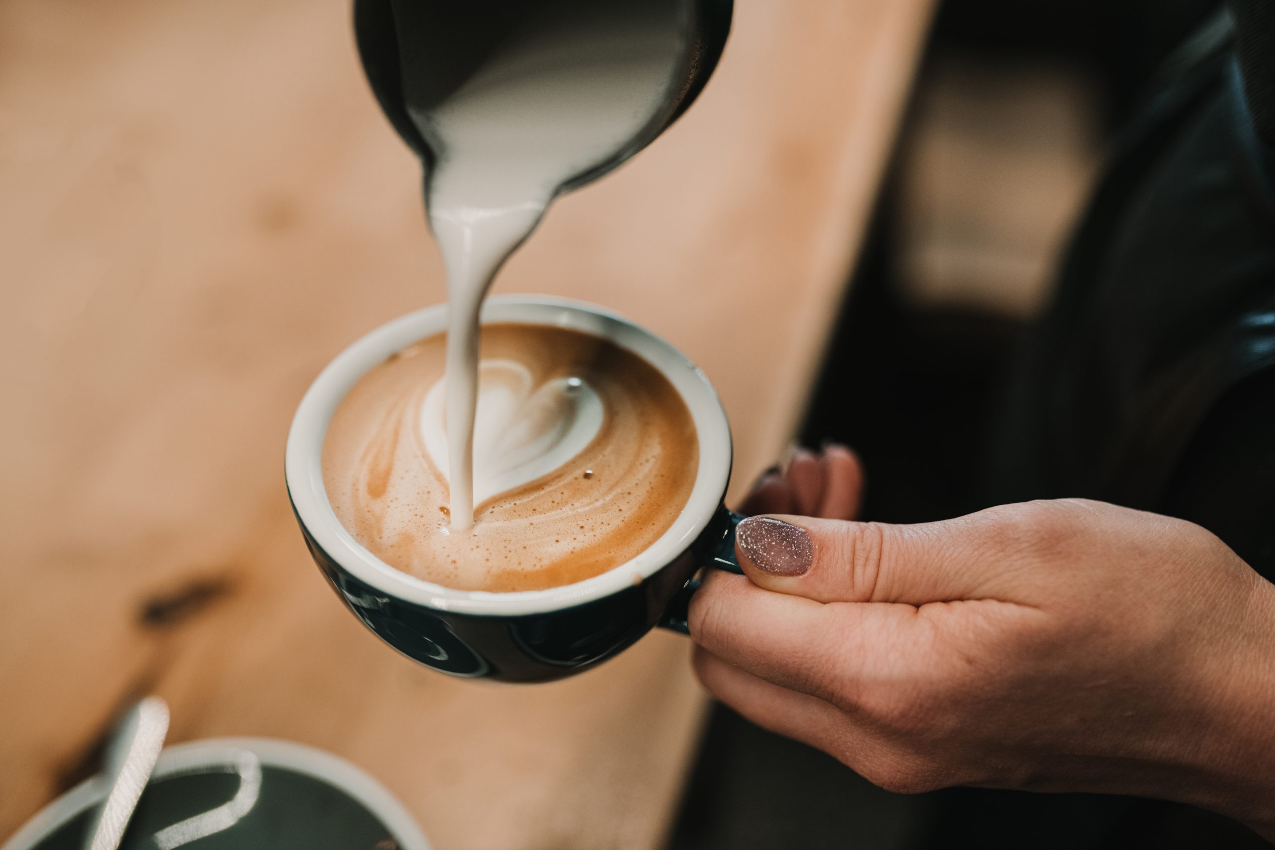 Steamed milk being poured into a cup of espresso to form a heart shape on a wooden counter