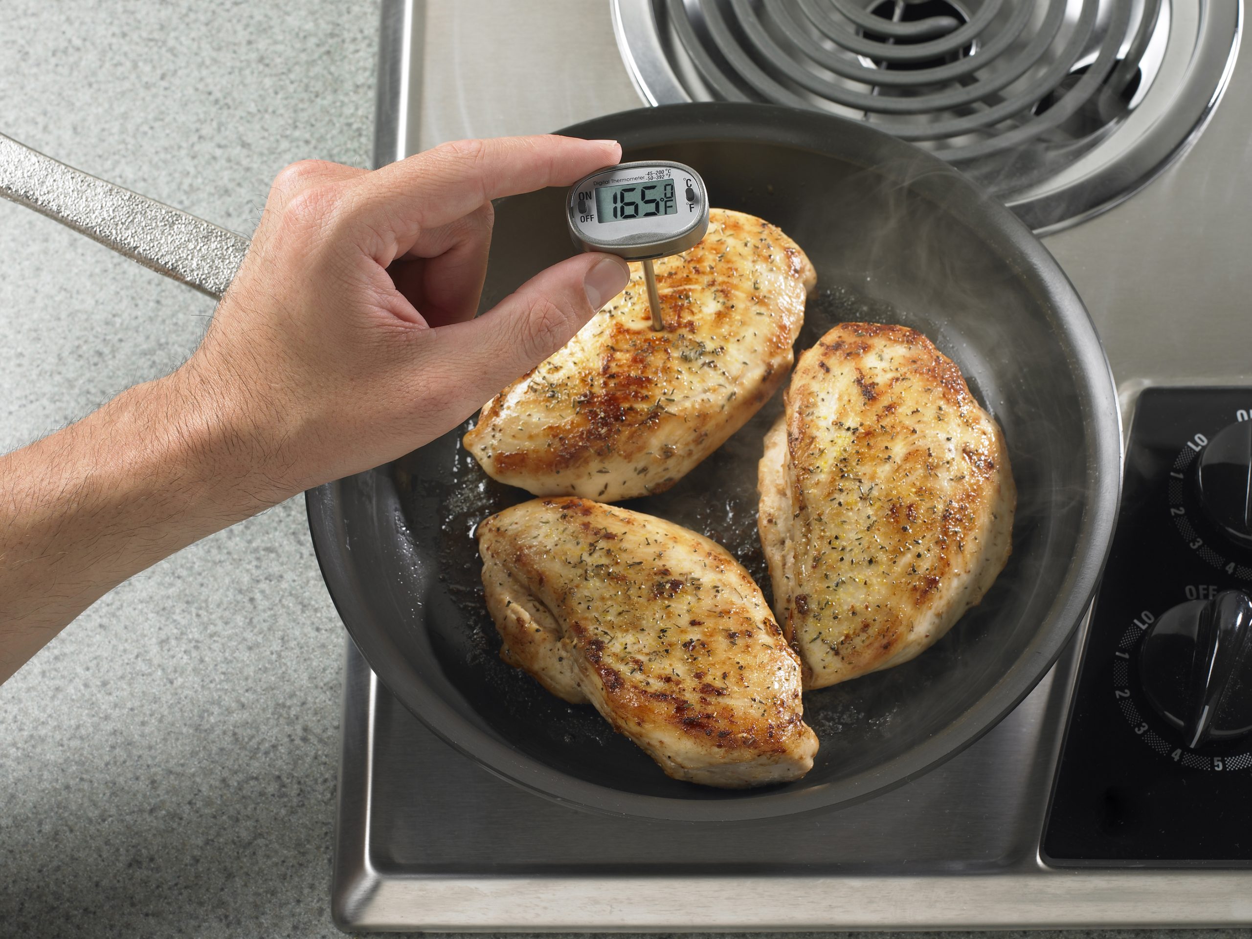 A hand holding a digital food thermometer in a pan of browned chicken breasts on a stovetop