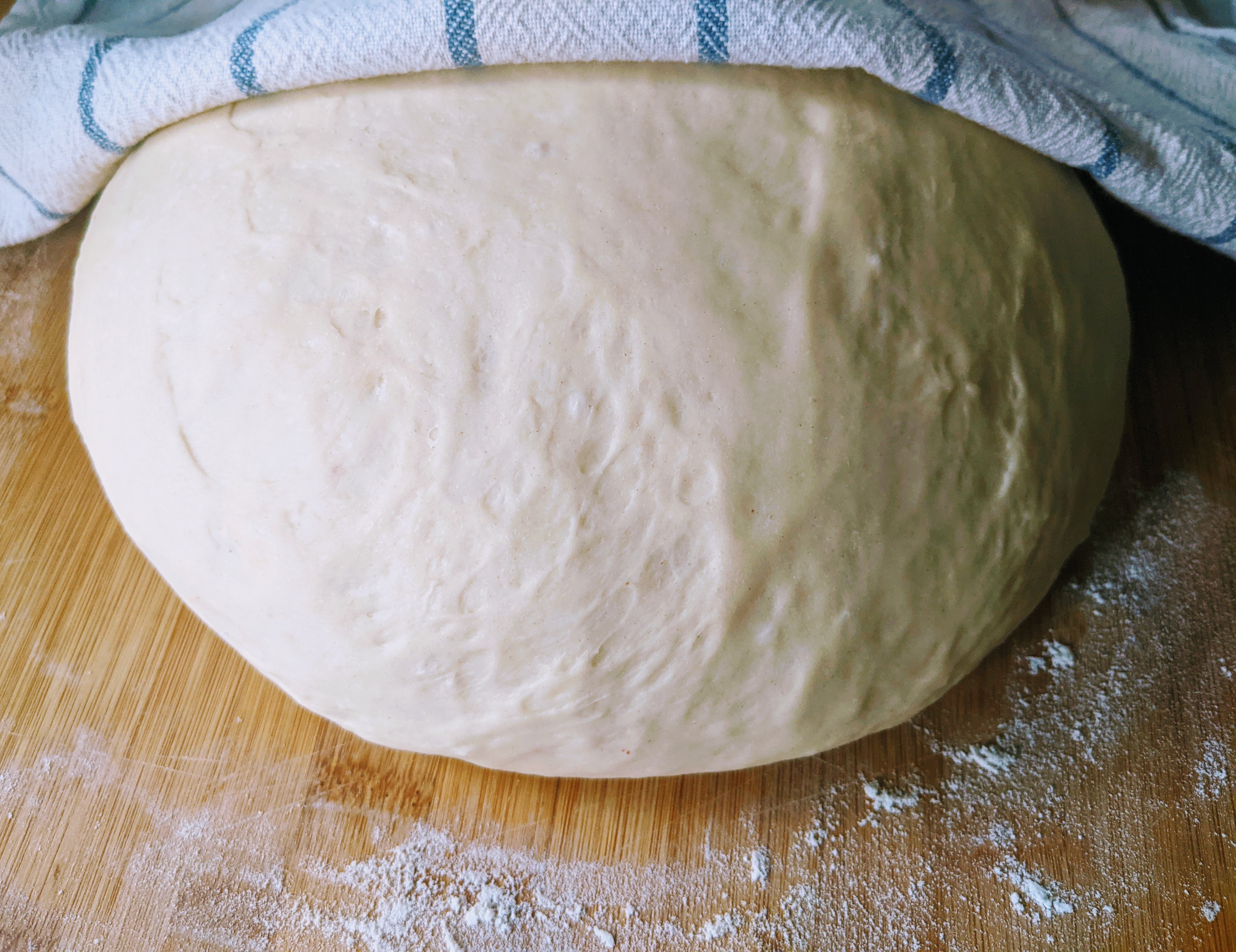 A smooth ball of risen raw bread dough on a floured wooden board under a striped cloth